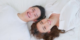 亚洲新婚夫妇躺在床上看着相机的肖像。漂亮漂亮的年轻男女穿着睡衣在家里的卧室里享受清晨的活动。家庭关系的概念