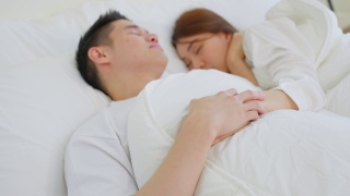 亚洲有吸引力的年轻夫妇躺在床上睡觉在卧室。美丽的新婚夫妇，穿着睡衣的男人和女人，清晨在房子里一起躺在舒适的枕头上，感到幸福和放松。视频素材模板下载