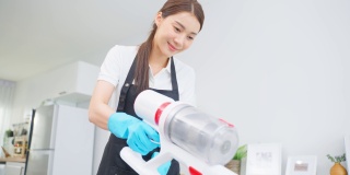 亚洲清洁服务女工清洁客厅在家里。美丽快乐的女孩，家庭主妇管家清洁工穿围裙和吸尘凌乱肮脏的桌子家务或家务。