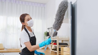 亚洲清洁服务女工清洁厨房和家庭。漂亮的女管家清洁工戴着口罩，用鸡毛掸子擦拭凌乱的柜台，为管家做家务或杂务。视频素材模板下载