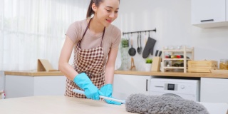 亚洲清洁服务女工清洁厨房和家庭。年轻漂亮的女管家清洁工穿着围裙，用鸡毛掸子擦拭餐桌，为家务或杂务家务做家务。