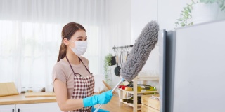 亚洲清洁服务女工清洁厨房和家庭。漂亮的女管家清洁工戴着口罩，用鸡毛掸子擦拭凌乱的柜台，为管家做家务或杂务。