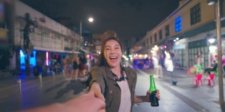亚洲女游客的肖像，牵着男友的手，走在街上。在城市旅游的年轻男女花时间在一起度假，在漆黑的夜晚在路上喝一瓶啤酒。