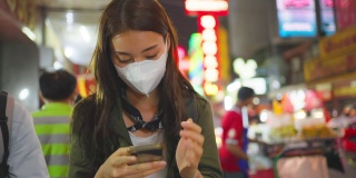 亚洲有吸引力的女人背包客聊天电话，而走在城市。美丽的女游客在街上独自旅行时戴着口罩，在夜间用智能手机与朋友交流。