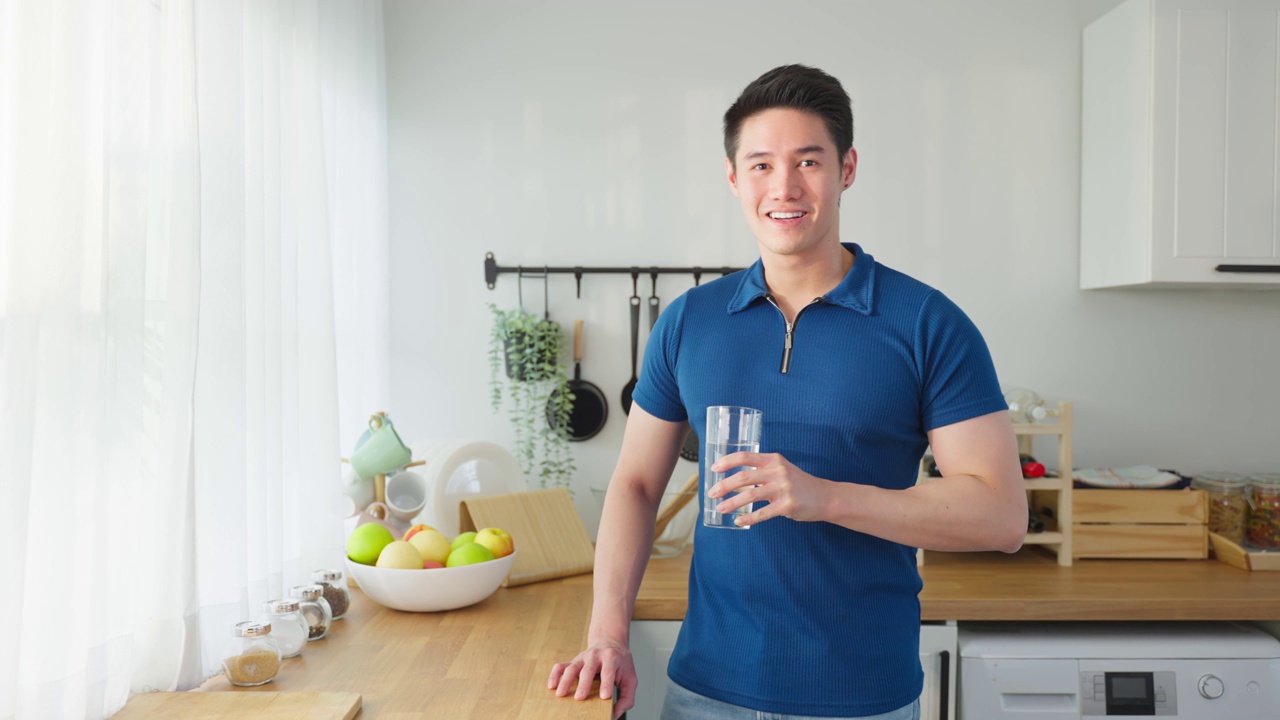 亚洲有吸引力的男性的肖像，在厨房里拿着一杯水。年轻饥渴的帅哥在清晨醒来后，为居家保健而斟上一杯纯净的天然矿物质
