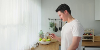 亚洲帅哥站在家里厨房里打电话。快乐有魅力的男性早上站在窗外享受假日周末，用智能手机与科技交流