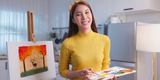 亚洲年轻艺术家女孩在画板上色的肖像在家里。有吸引力的女性绘画艺术图片，创造艺术品与水彩和画笔享受创意活动和看着相机。