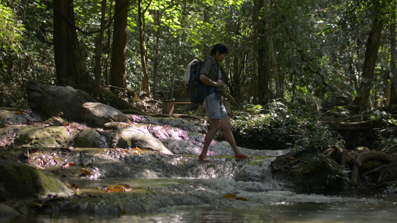 夏季，亚洲女性徒步旅行者背着双肩包，在美丽的热带森林中穿过流过岩石的溪流。