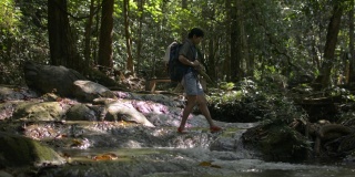 夏季，亚洲女性徒步旅行者背着双肩包，在美丽的热带森林中穿过流过岩石的溪流。