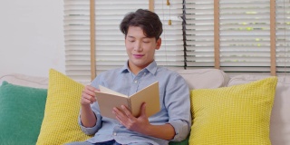 快乐英俊的亚洲男人坐在沙发上微笑和阅读书籍放松时间在家里。兴高采烈的商人穿着蓝色衬衫，坐在沙发上看书