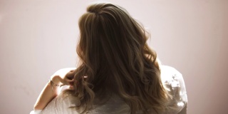 一个年轻的女人在米色的墙壁背景上摇着她金色的卷发。艺术。后视图，一个年轻的金发女人在一个白色的衬衫摆姿势和触摸她的头发。