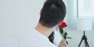 亚洲青年惊喜送玫瑰给美丽的女友。迷人浪漫的新婚男女坐在床上，一起享受庆祝周年纪念日和情人节