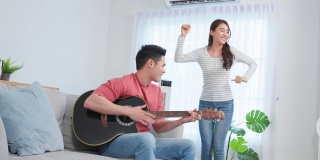亚洲年轻情侣在家里的客厅里一起弹吉他。迷人浪漫的新婚，活跃浪漫的男士坐在沙发上弹着乐器与美丽的女友共舞。