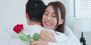 亚洲青年惊喜送玫瑰给美丽的女友。迷人浪漫的新婚男女坐在床上，一起享受庆祝周年纪念日和情人节