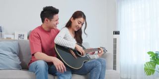 亚洲年轻情侣在家里的客厅里一起弹吉他。迷人浪漫的新婚，活跃浪漫的男人坐在沙发上教漂亮的女朋友在家里弹琴。