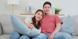 亚洲年轻情侣在家中客厅一起看电影。有吸引力的浪漫的新婚姻，男人和女人坐在沙发上，看电视节目，有乐趣的笑在一起的房子。