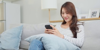 亚洲美女坐在沙发上，在家里用手机聊天。快乐迷人的年轻女孩在家里度过休闲时光，放松心情，享受在客厅用智能手机交流和讨论。