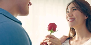 亚洲青年惊喜送玫瑰给美丽的女友。迷人浪漫的新婚男女花时间在房子里一起庆祝周年纪念日和情人节。