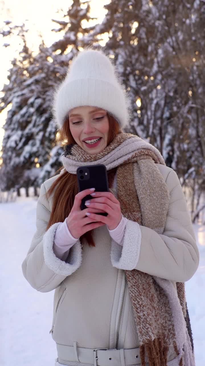 垂直拍摄快乐的年轻女子穿着温暖的冬装，用智能手机拍摄城市公园的冬季风景。