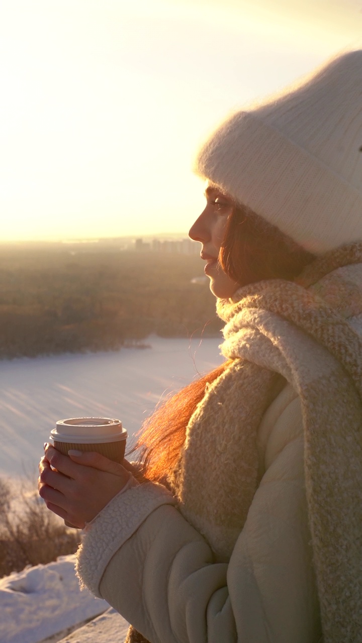 在美丽的冬季雪景的背景下，有吸引力的年轻女子看着日落和喝热咖啡的垂直肖像。