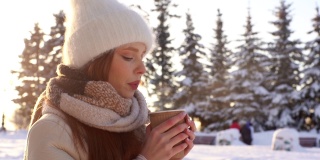 在雪城公园里，迷人的年轻女子戴着白色帽子，闭着眼睛享受着从杯子里拿出来的热的芳香咖啡或巧克力。