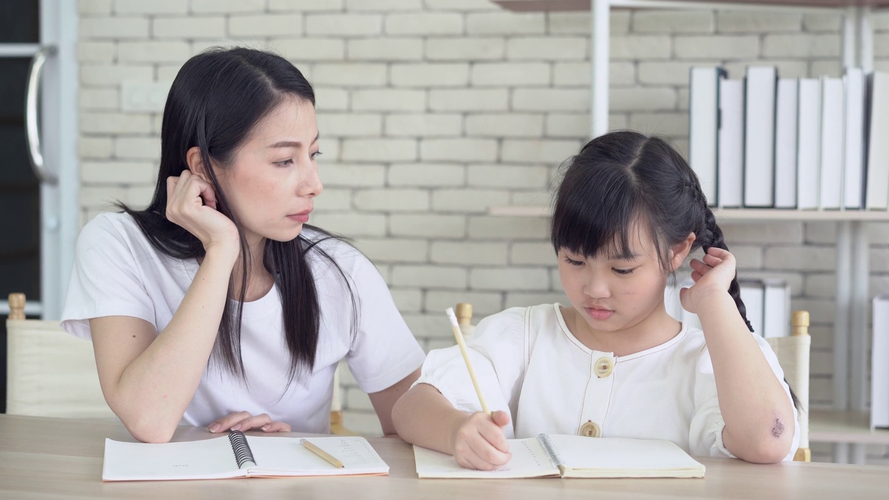 愤怒的年轻亚洲母亲生气的小女儿教和做作业写。她坐在家里的书桌前听不懂。孩子的教育问题，父母和孩子的冲突。