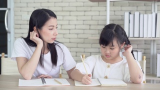 愤怒的年轻亚洲母亲生气的小女儿教和做作业写。她坐在家里的书桌前听不懂。孩子的教育问题，父母和孩子的冲突。视频素材模板下载