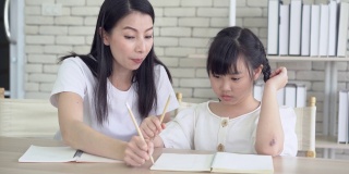 精疲力竭的年轻亚洲单身母亲头痛，教小女儿做作业。小女孩不懂家庭作业，她妈妈对此很不高兴。孩子的教育问题。