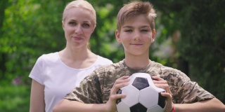 积极的十几岁的男孩摆姿势与足球，模糊的军事妇女把手放在肩膀上的慢动作的肖像。快乐自信的白人儿子和骄傲的母亲在公园摆姿势。