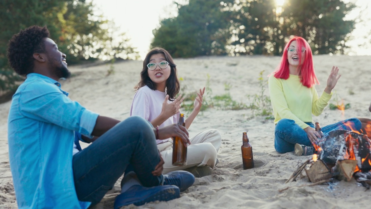 朋友们坐在湖边的沙滩上，一起消磨时间，在篝火旁玩耍，放松，一个女人弹吉他，唱歌，喝啤酒