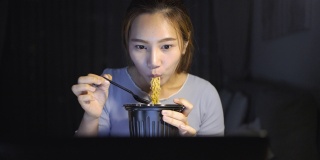 亚洲自由职业者聪明的商业妇女吃方便面，而工作在家里的客厅在笔记本电脑在晚上快乐的亚洲女孩坐在桌子上享受放松的时间