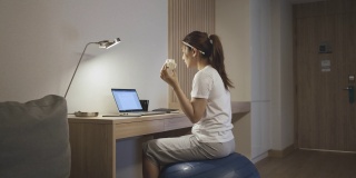 亚洲女性在瑜伽球上放松，吃三明治，和同事们在笔记本电脑上聊天，晚上在家工作。