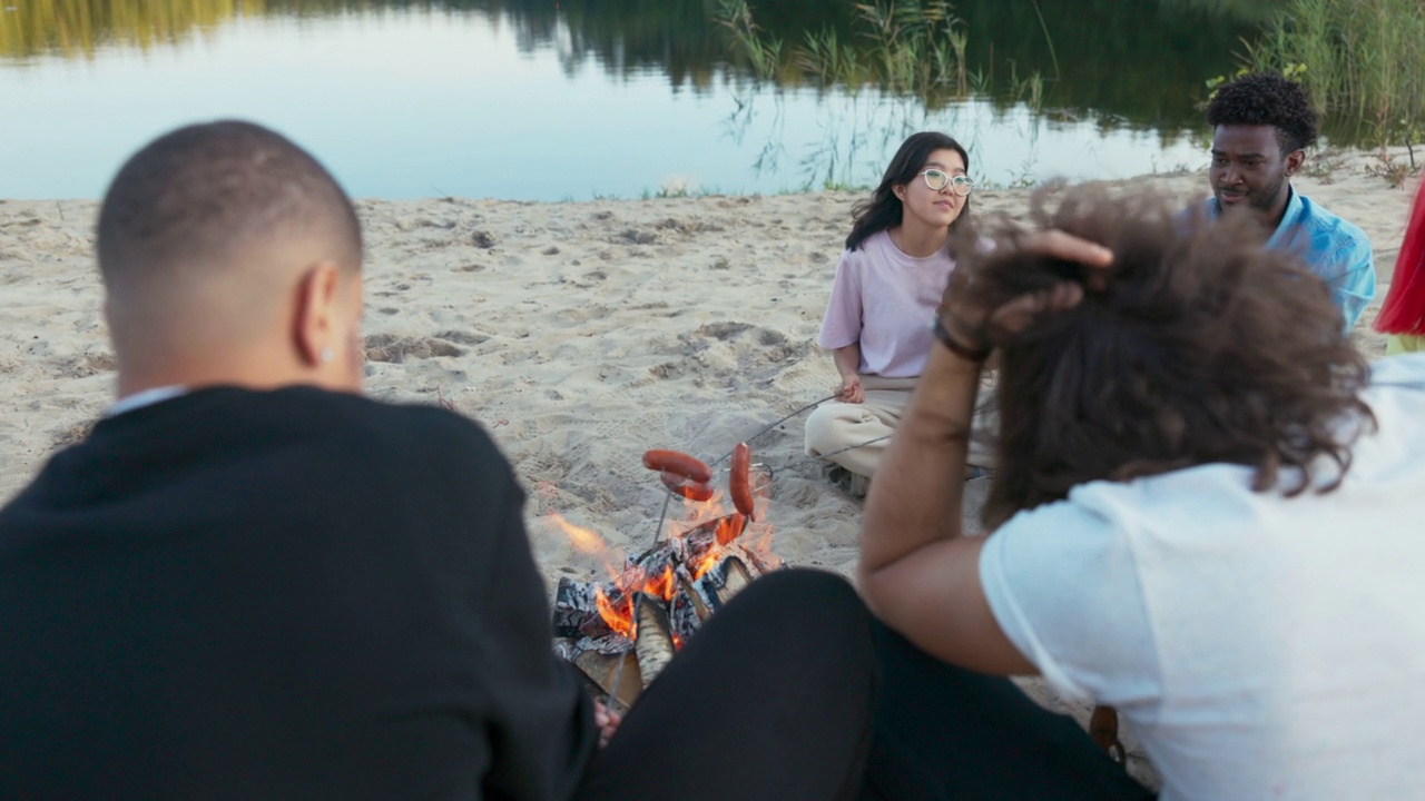 戴着眼镜的美女坐在湖边的沙滩上，拿着一根香肠棒在篝火上烤，把香肠棍两面都炸得粉碎，和朋友们在一起消磨时光