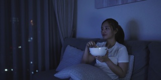 亚洲妇女看电视悬疑电影或新闻看起来快乐和有趣的和吃爆米花在家庭隔离时间在家里客厅沙发深夜。