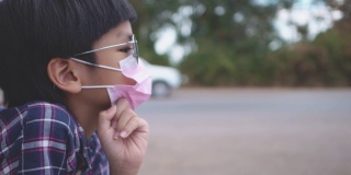 小亚洲男孩戴着面具和太阳镜坐在路边，而汽车经过。