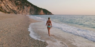 空中低空跟随成年妇女享受美丽的米洛斯海滩的Lefkada，爱奥尼亚岛，希腊。夕阳金色的阳光和原始的海浪打破了蓝绿色的大海在岸边