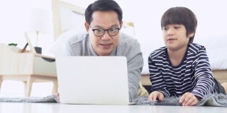 愉快的亚洲家庭一起在房间里度过时光。快乐的亚洲父亲和儿子躺在地毯上，在家里的卧室里使用笔记本电脑。