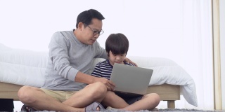 一位亚洲父亲坐在家里卧室的地板上，一边用笔记本电脑做作业，一边和可爱的儿子聊天。在家学习和远程学习，在线教育的概念。