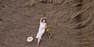 穿着漂亮衣服的年轻女子躺在麦田里。暑假的概念。快乐的女孩戴着柳条帽，手拿包，带着向日葵在大自然上。4 k