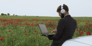 一个商人戴着耳机，穿着西装，在他的汽车引擎盖上用笔记本电脑工作在绿色的田野里，红色的郁金香。
