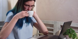 一位蓝色头发、戴着眼镜、疲惫不堪的女学生正在笔记本电脑上打字，一边喝着咖啡，一边打着哈欠。女孩坐在电脑前睡着了。工作中的倦怠概念
