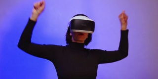 快乐女孩在虚拟现实头盔举起双手，跳舞，紫色霓虹灯，全高清。虚拟世界的概念。虚拟空间的生活方式