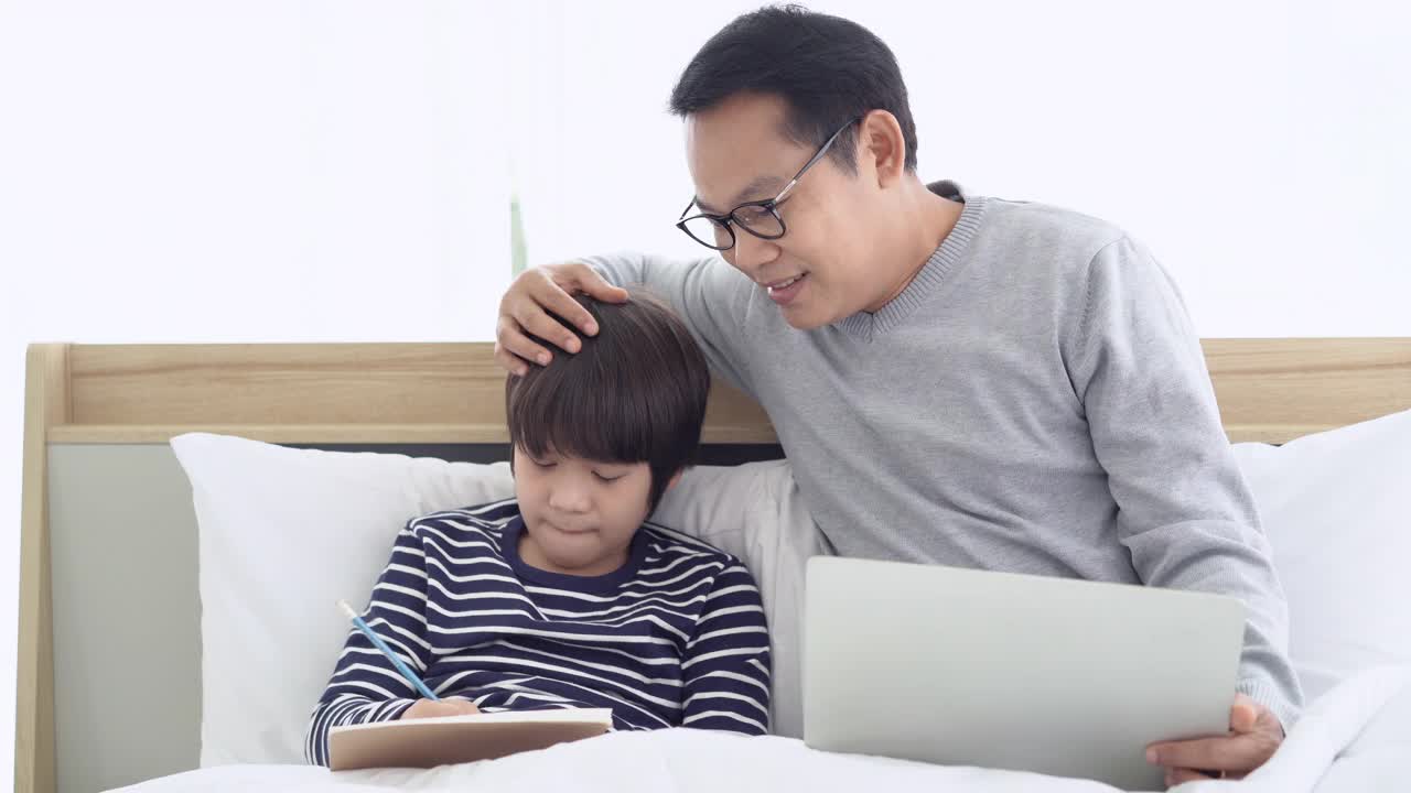 居家办公快乐的亚洲父亲使用笔记本电脑，和儿子聊天，同时做作业，坐在床上的卧室在家里。自我隔离