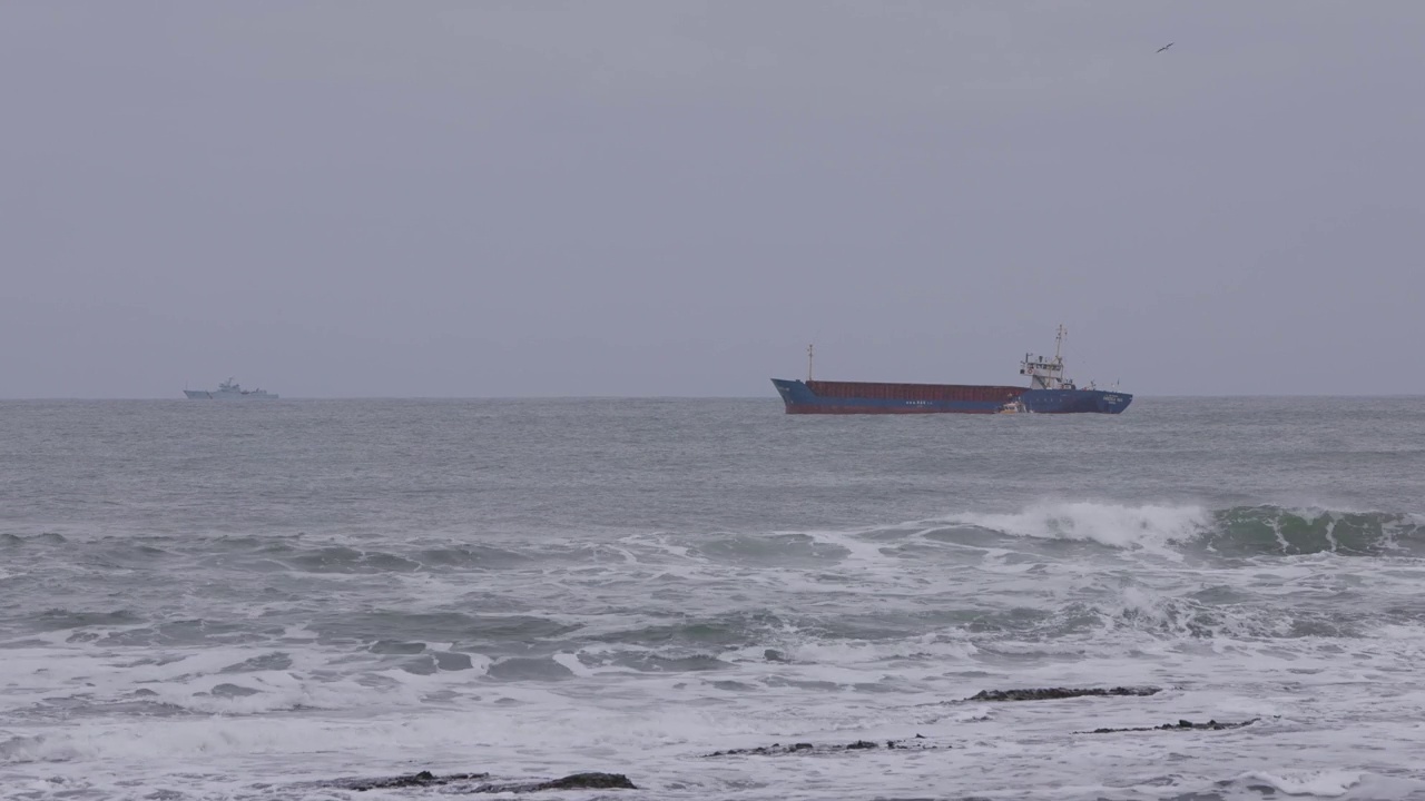 在暴风雨天气下波涛汹涌的海面上，一艘油轮沿着一艘领航船和一艘军用船的镜头在背景中出现