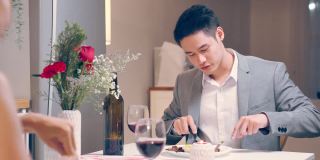 亚洲年轻情侣共进晚餐庆祝情人节。迷人浪漫的新婚男女在家里的厨房里举行晚会，在餐桌上吃结婚纪念日的食物。