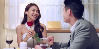亚洲青年惊喜送玫瑰给美丽的女友。迷人浪漫的新婚夫妇，男人和女人一起吃晚餐，庆祝周年纪念日和情人节在家里