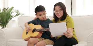 亚洲母亲和儿子坐在房子的白色沙发上儿子弹吉他，母亲唱歌