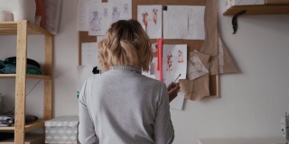 女时装设计师看着钉在她桌子后面墙上的图纸和草图。工作室是阳光明媚的。