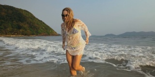 海边的沙滩上，穿着白色束腰外衣的女人。金发碧眼的太阳镜。享受假期的游客，在水上跑步，散步很有趣