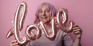 情人节。漂亮年轻的30多岁的女人，在粉红色的背景下，拿着粉红色的箔纸爱情词气球玩得很开心。母亲节,生日。